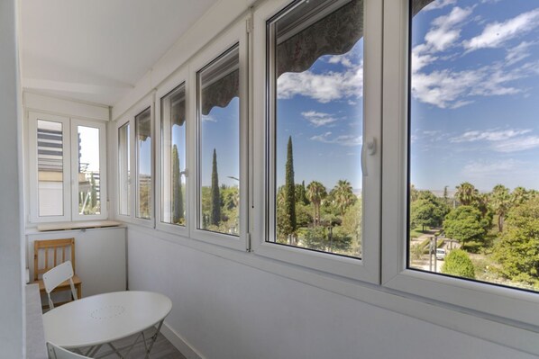 A room with a white table and chair, large windows with decorative shutters, and a view of trees and a clear sky.