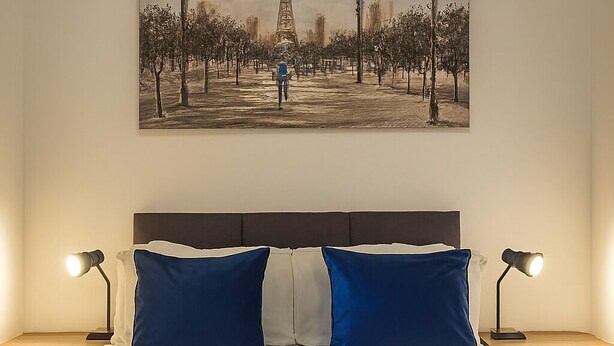A neatly made bed with two pillows, two bedside lamps, and a folded towel on each side. - The Nivora Suite (Newsome)