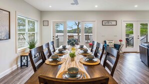 A dining room with a wooden table set for six, surrounded by chairs, with a ceiling fan above and a view of the outdoors through large windows.