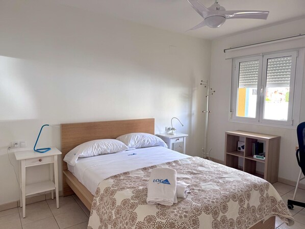 A hotel room with a bed, a desk, a chair, a ceiling fan, a window with blinds, and a small shelf with books.