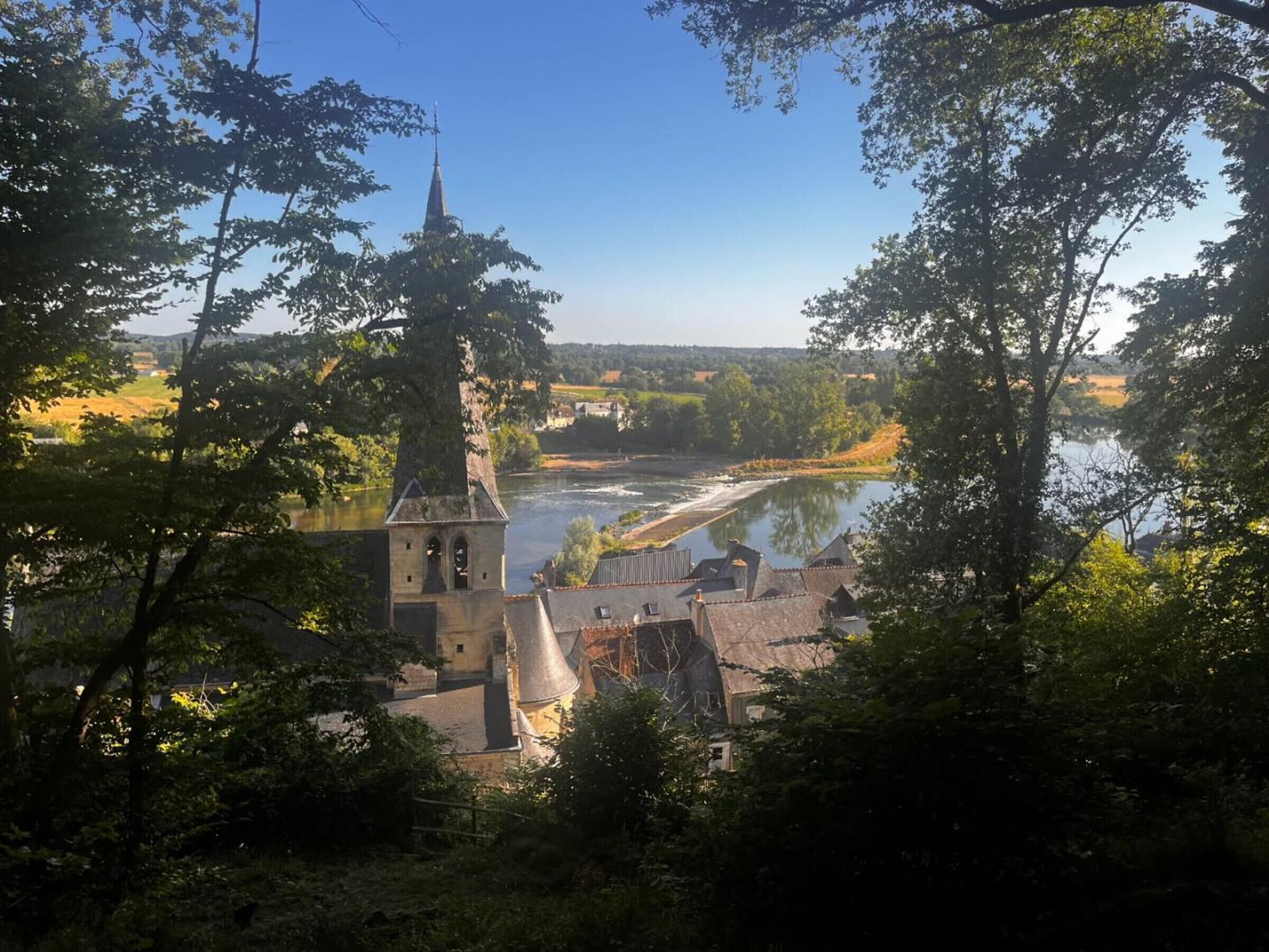 Un bâtiment historique doté d’une tour et d’une cloche imposantes, entouré d’une végétation luxuriante et bordé par une rivière.