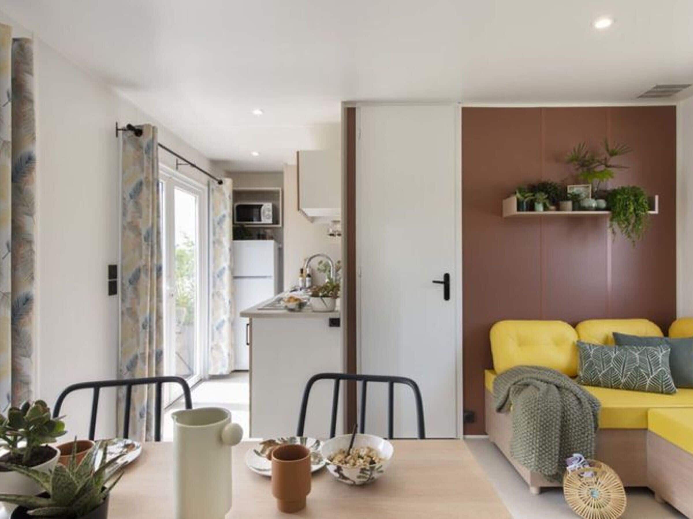 A modern living space with a dining area, kitchen, and a yellow sofa with patterned cushions.