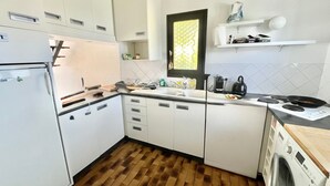 Une cuisine compacte avec des meubles blancs, un lave-linge encastré et une fenêtre agrémentée d’un store à motifs.