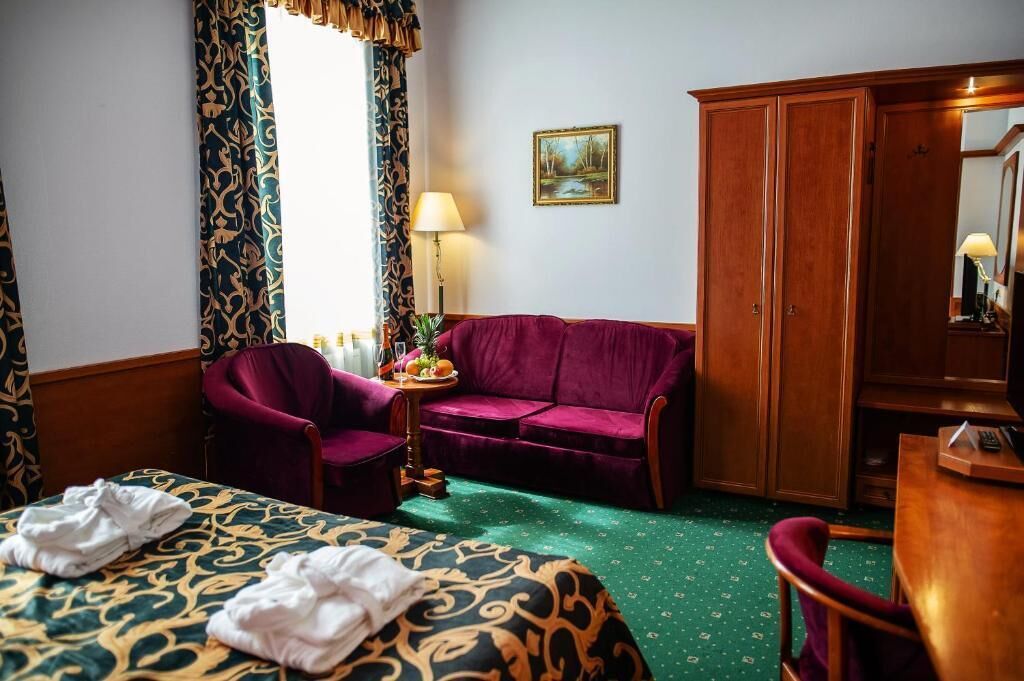 Habitación de hotel con cama, sofá púrpura, armario de madera y una pequeña mesa con lámpara y fruta.