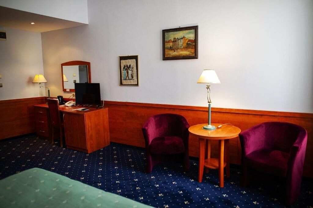 Une chambre d’hôtel équipée d’un bureau, de deux chaises, d’un lit, d’une lampe de bureau et de deux tableaux accrochés au mur.