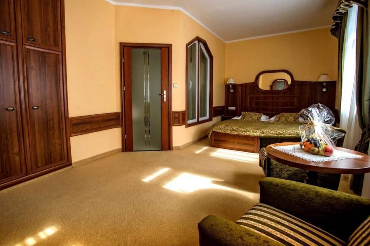 Une chambre d’hôtel avec un lit, une armoire en bois, un miroir, une petite table ronde avec un panier de fruits et un fauteuil vert.