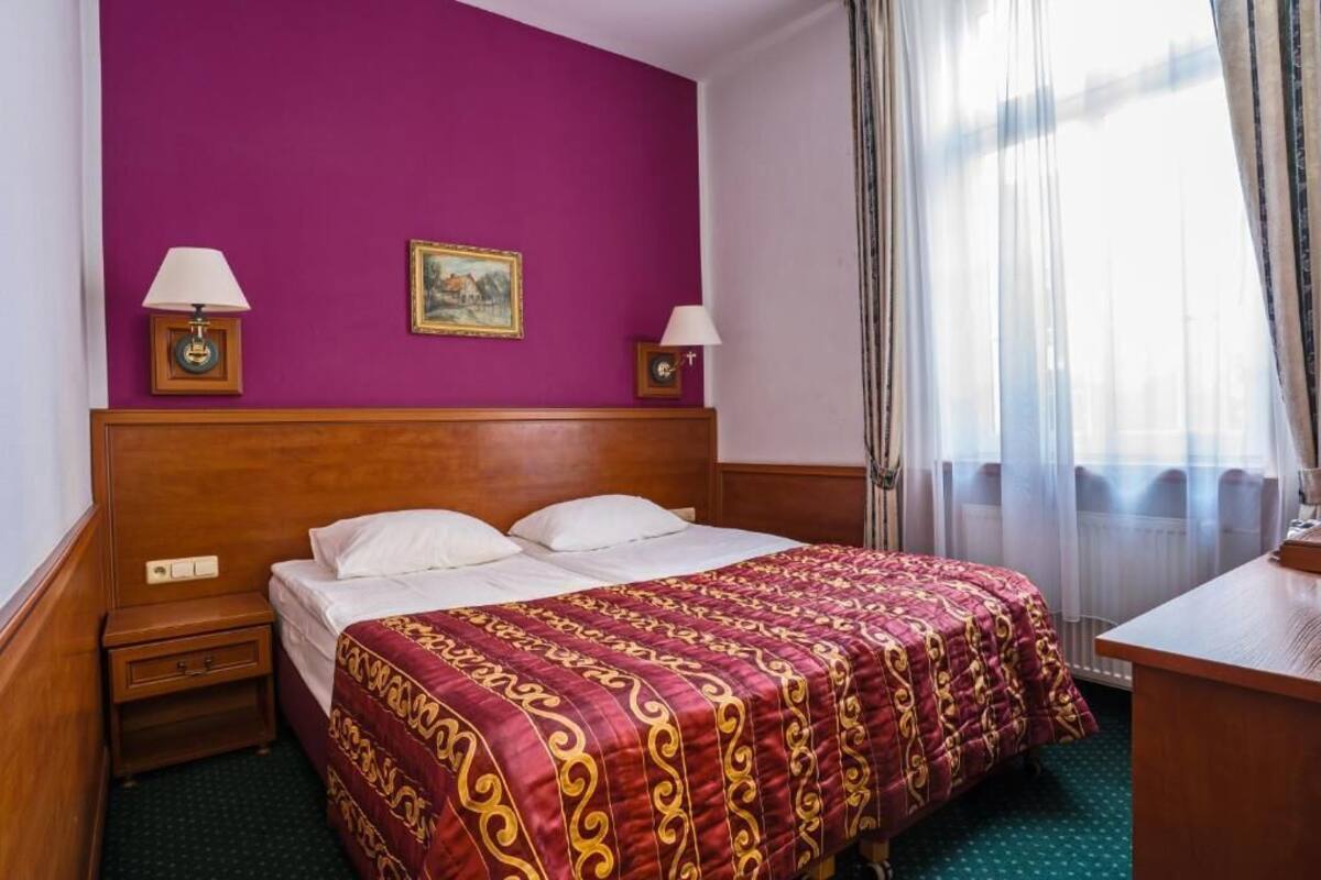Une chambre d’hôtel avec une tête de lit en bois, un grand lit recouvert d’une couvre-lit à motifs, deux lampes de chevet, une petite table et une fenêtre ornée de rideaux légers.