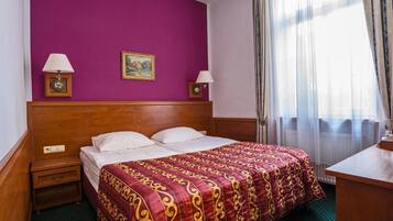 Habitación de hotel con cabecera de madera, cama grande con colcha estampada, dos lámparas de noche, una mesita y una ventana con cortinas de encaje.