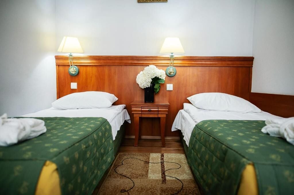 Habitación de hotel con dos camas, cada una con cubrecama verde y almohadas blancas. Hay una mesita de noche con una lámpara entre las camas, y un florero con flores blancas sobre la mesita.