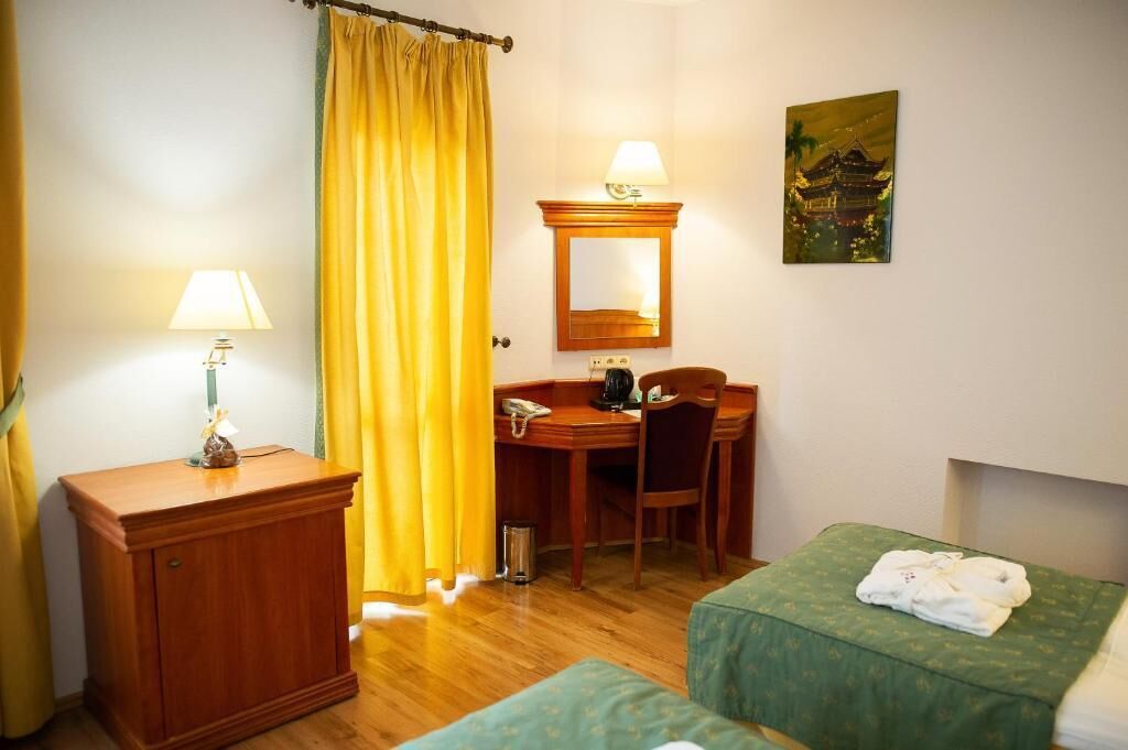 A hotel room with a wooden desk, a chair, a small cabinet, a bed with a green bedspread, and a painting on the wall.