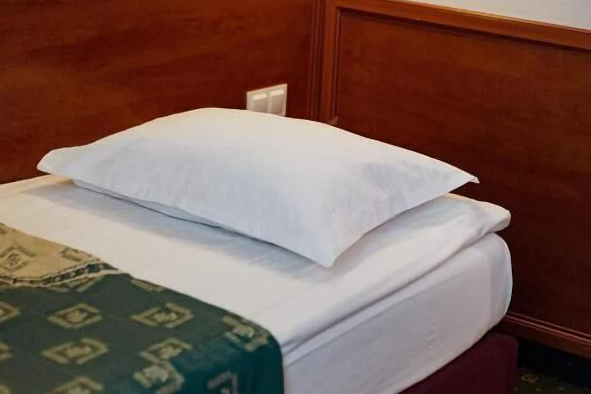 Un lit bien fait, avec du linge de lit blanc et une couverture à motifs verts, une tête de lit en bois et une lampe fixée au mur.