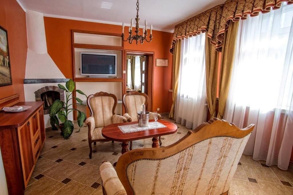 A room with a wooden dining table, two upholstered chairs, a chandelier, a television, and a large window with curtains.