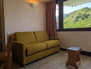 A room with a sofa, a small table, a window with a view of a hill, and a patterned curtain.