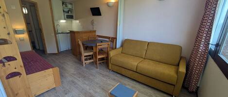 Un espace de vie compact comprenant un canapé, une petite table, un banc en bois et, au fond, une kitchenette.