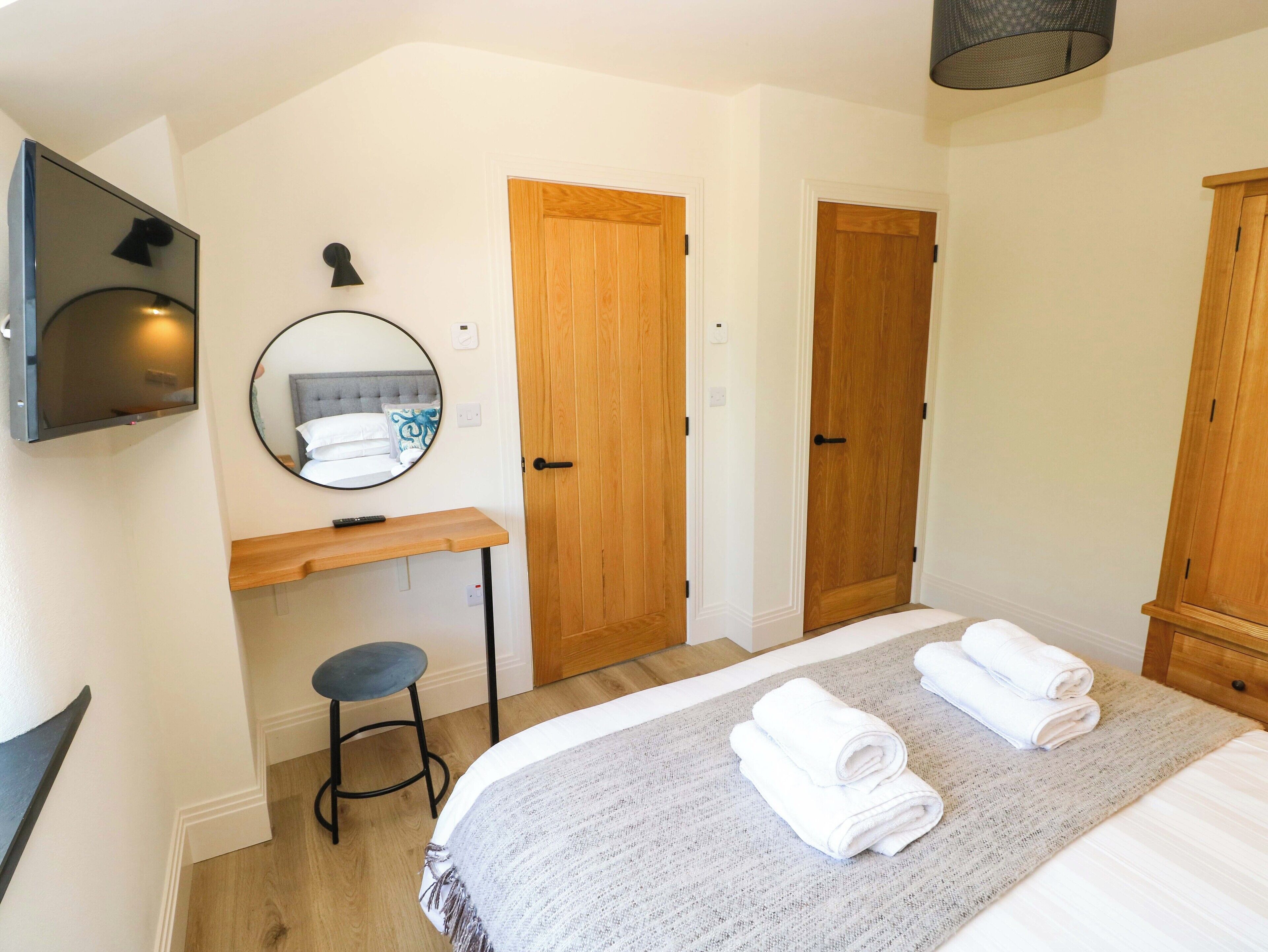 Une chambre d’hôtel avec un lit, deux portes en bois, un miroir rond, une télévision, un tabouret et un petit bureau.