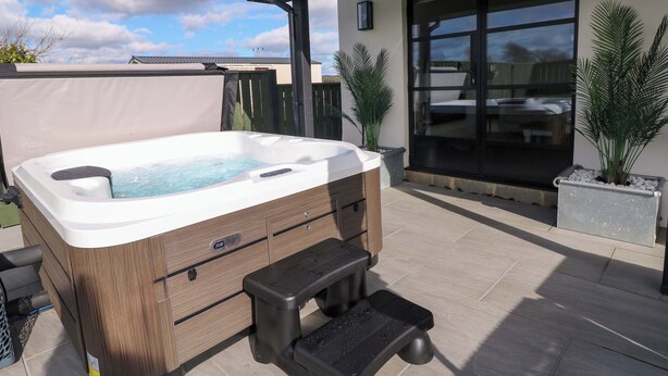 Un jacuzzi con revestimiento de madera y un taburete, situado en un patio con suelo de baldosas y plantas en macetas.
