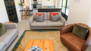 Una sala de estar con un sofá, un sillón de cuero marrón y una mesa de centro de madera. Se observa una cocina al fondo, con un televisor y una planta.