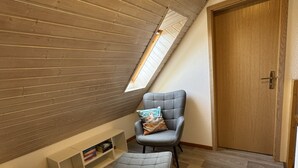 Camera soppalcata con soffitto spiovente, un lucernario, una poltrona grigia con cuscino, un poggiapiedi e una porta in legno.