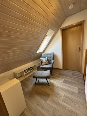 A loft bedroom with a sloped ceiling, a skylight, a grey armchair with a cushion, a small ottoman, and a wooden door. - Detached vacation home in Nessmersiel (near Deich): own garden & wallbox (Dornum)