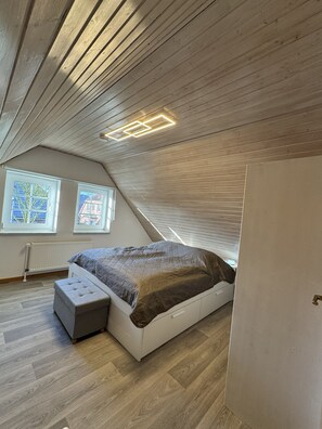 A bedroom with a sloped ceiling, a bed with a storage bench, and two windows with views of buildings. - Detached vacation home in Nessmersiel (near Deich): own garden & wallbox (Dornum)
