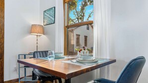 A dining area with a wooden table, black chairs, a lamp, and a window with a view of trees and buildings. - Secret Garden Cottage - Walk to Main St. (Fredericksburg)