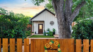 A wooden fence with a sunflower design, a house with a white exterior, a brown door, and a tree in front. - Secret Garden Cottage - Walk to Main St. (Fredericksburg)