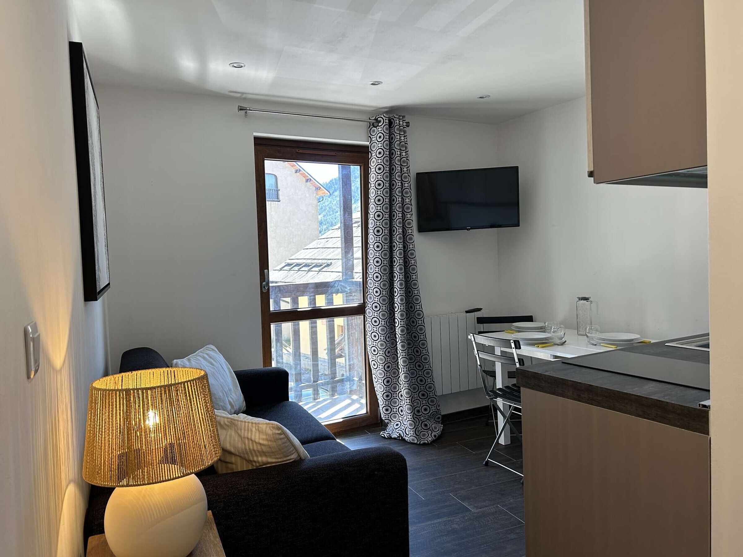 En kompakt opholdsstue med sofa, spisebord, tv ophængt på væggen og et vindue med udsigt til en bygning.