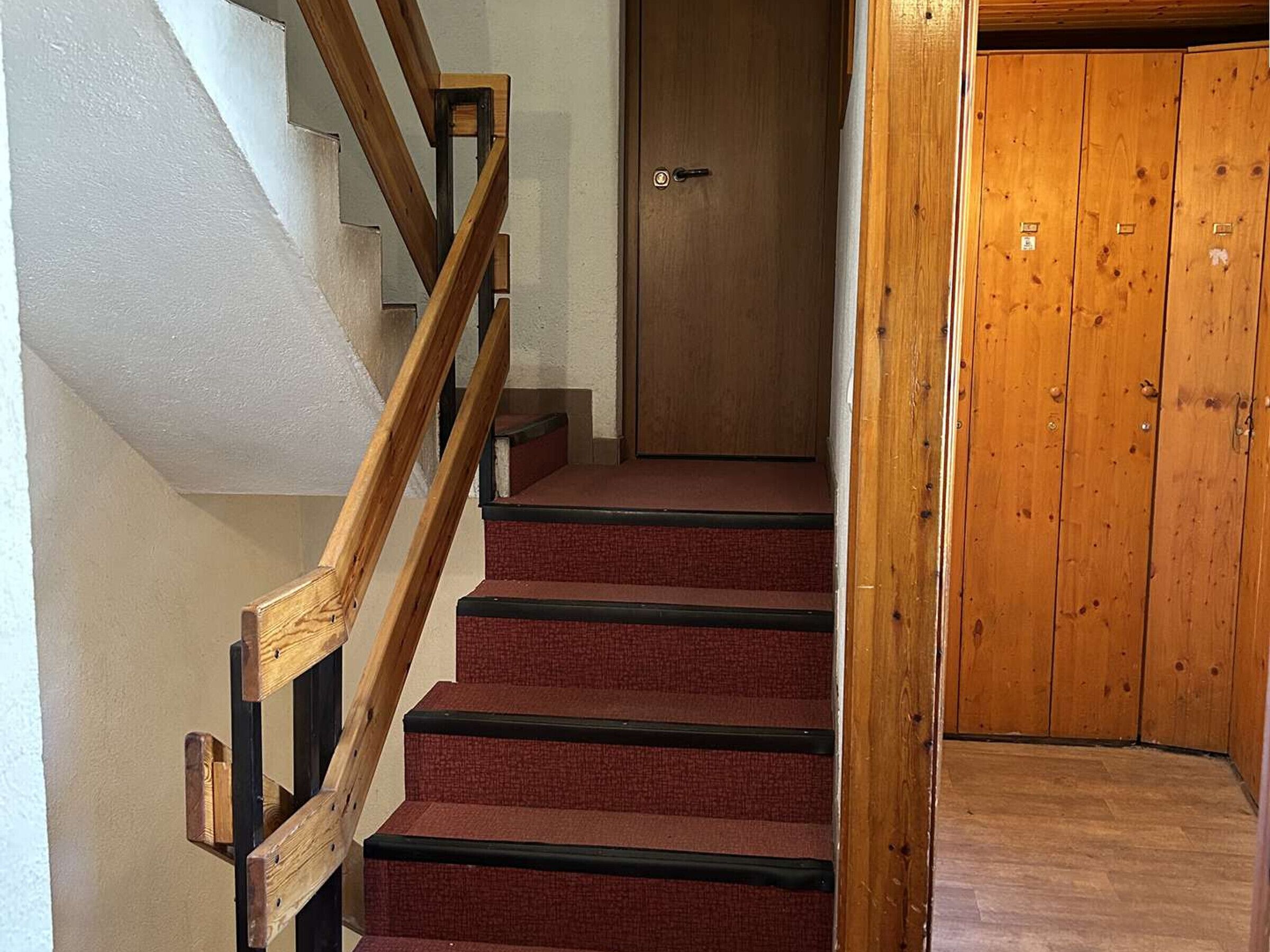 Un escalier avec des rampes en bois et des marches recouvertes d’une moquette rouge, menant à une porte en bois.