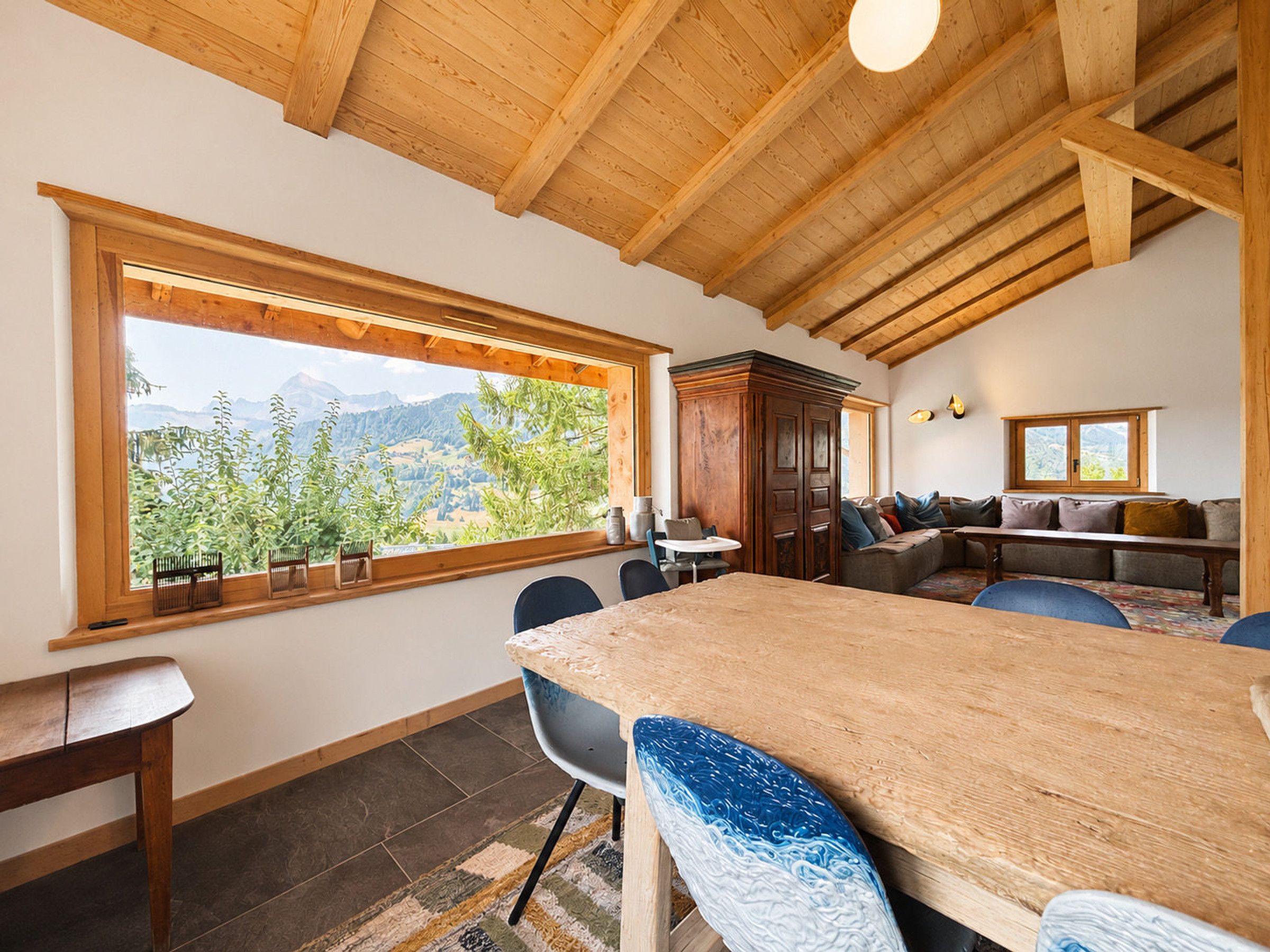 A dining area with a wooden table and chairs, a large window with a mountain view, and a wooden ceiling.