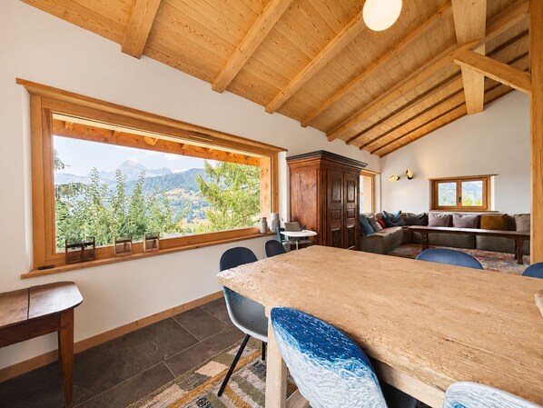 Een eetgedeelte met een houten tafel en stoelen, een groot raam met uitzicht op bergen, en een houten plafond.