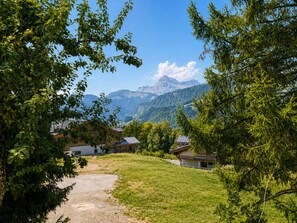 A rural landscape with a clear path leading to a cluster of traditional wooden houses, surrounded by lush greenery and mountains in the background.