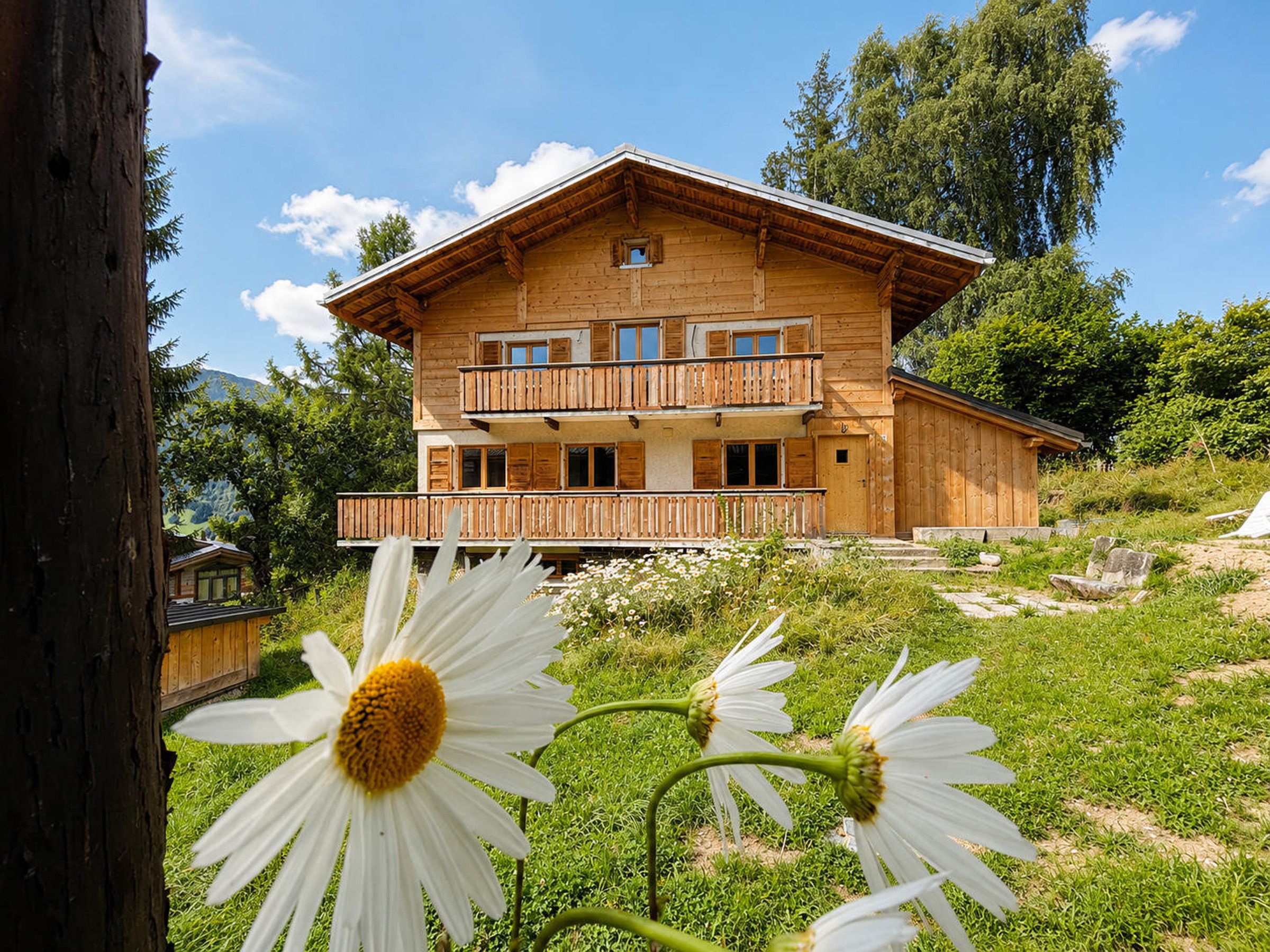 Un chalet en bois à deux étages, doté d’un balcon, entouré de verdure et de montagnes.