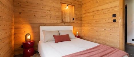 A wooden cabin room with a bed, two bedside lamps, a red rug, and a wooden ceiling.