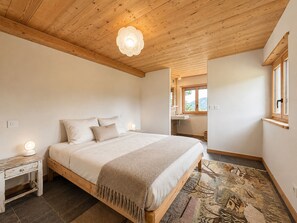 A bedroom with a wooden ceiling, a bed with a headboard, a nightstand, a window, and a bathroom visible in the background. - Spacious chalet 10 pers., near slopes and shops with WIFI (Notre-Dame-de-Bellecombe)