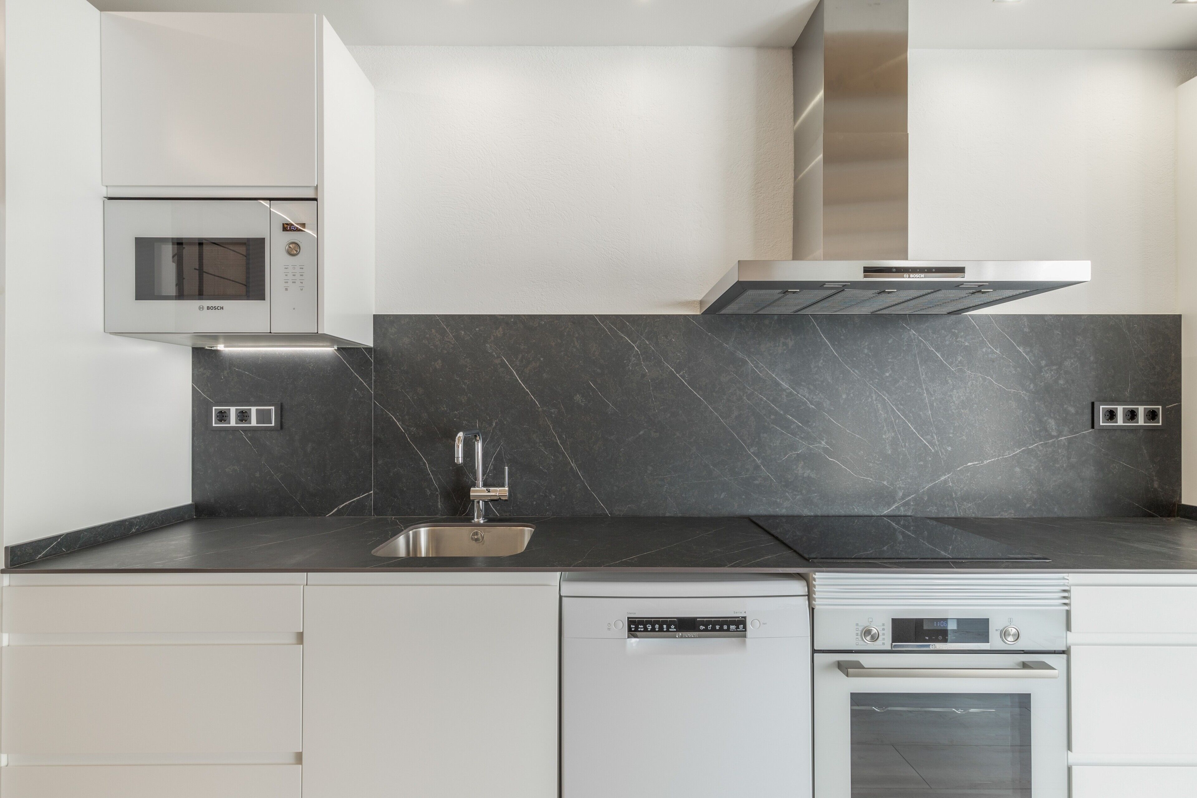 A modern kitchen with a Bosch microwave, dishwasher, and oven, a stainless steel sink, and a black marble backsplash.