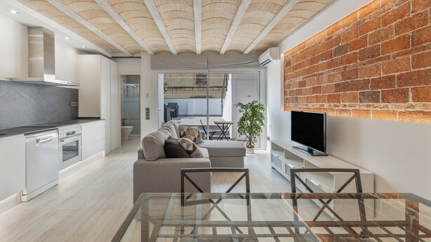 A modern interior with a kitchen, dining area, and living space featuring a brick wall, a flat-screen TV, and a glass table. - Holiday Home 'El Racó Del Mas' with Private Terrace, Wi-Fi & Air Conditioning (Canet de Mar)