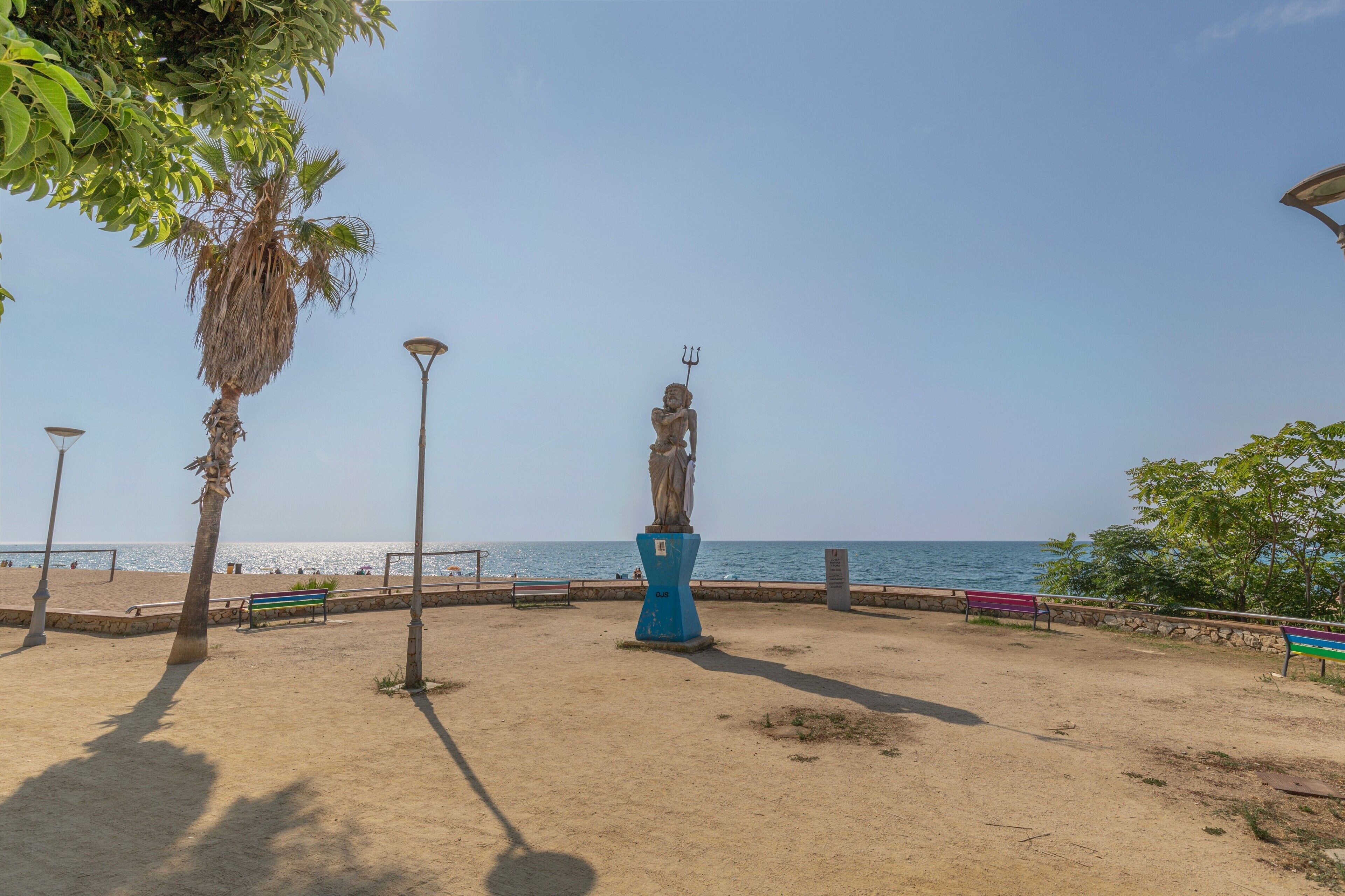 Een park aan het strand met een standbeeld, bankjes en een palmboom.