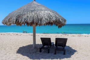 Une plage avec un parasol en chaume et deux chaises en bois.