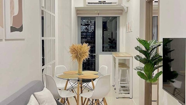 Una sala moderna con un sofá gris, una mesa de comedor de madera con sillas, una planta grande en maceta y un televisor de pantalla plana.
