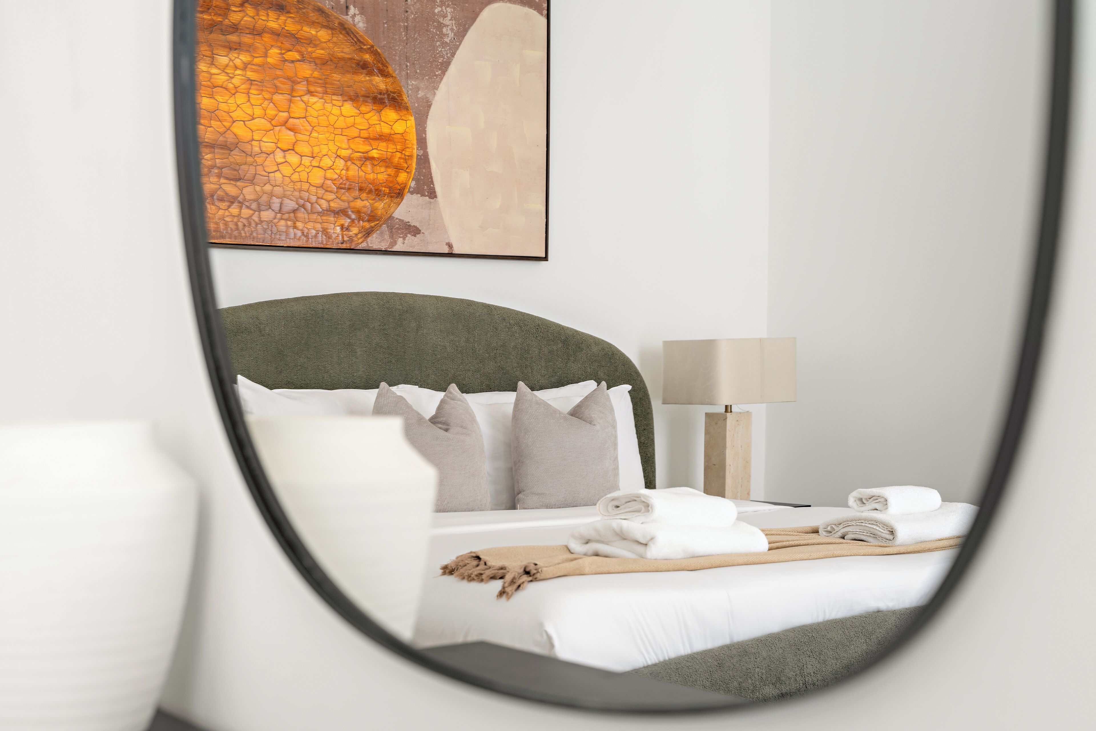 En pænt redt seng med hvide sengetæpper og en grøn sengegavl, en lampe på et natbord, og et indrammet kunstværk over sengen.