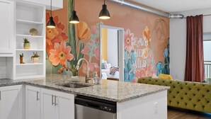 Une cuisine moderne avec un comptoir en granit, des armoires blanches et un lave-vaisselle. Un mur est décoré d’une fresque aux motifs floraux, et un canapé vert se trouve au fond de la pièce.