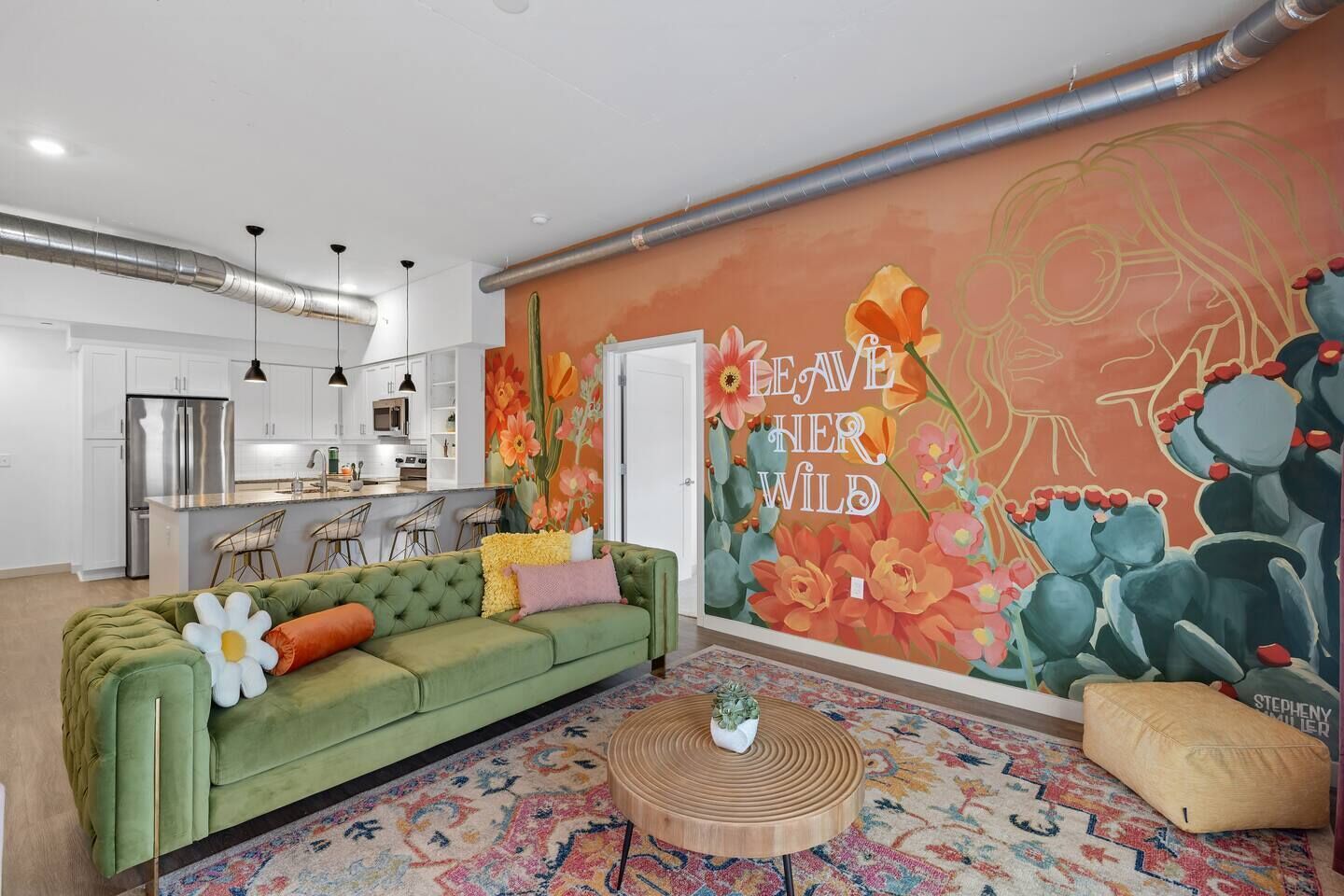 Una sala de estar moderna con un sofá verde, un tapete colorido y un mural que reza 'LEAVE HER WILD'.