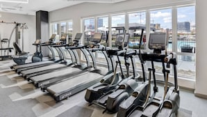 Une salle de sport moderne équipée de plusieurs tapis de course, de vélos d’appartement et d’un appareil d’aviron. De grandes fenêtres offrent une vue sur le paysage urbain et sur un plan d’eau.