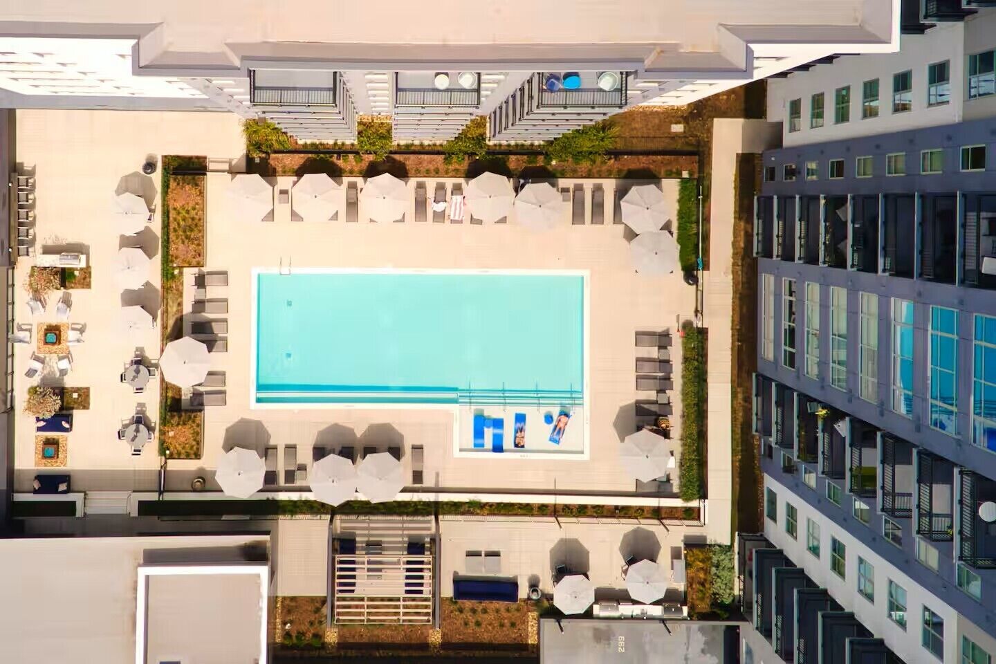 An aerial view of a rooftop pool area with lounge chairs and umbrellas, surrounded by buildings.