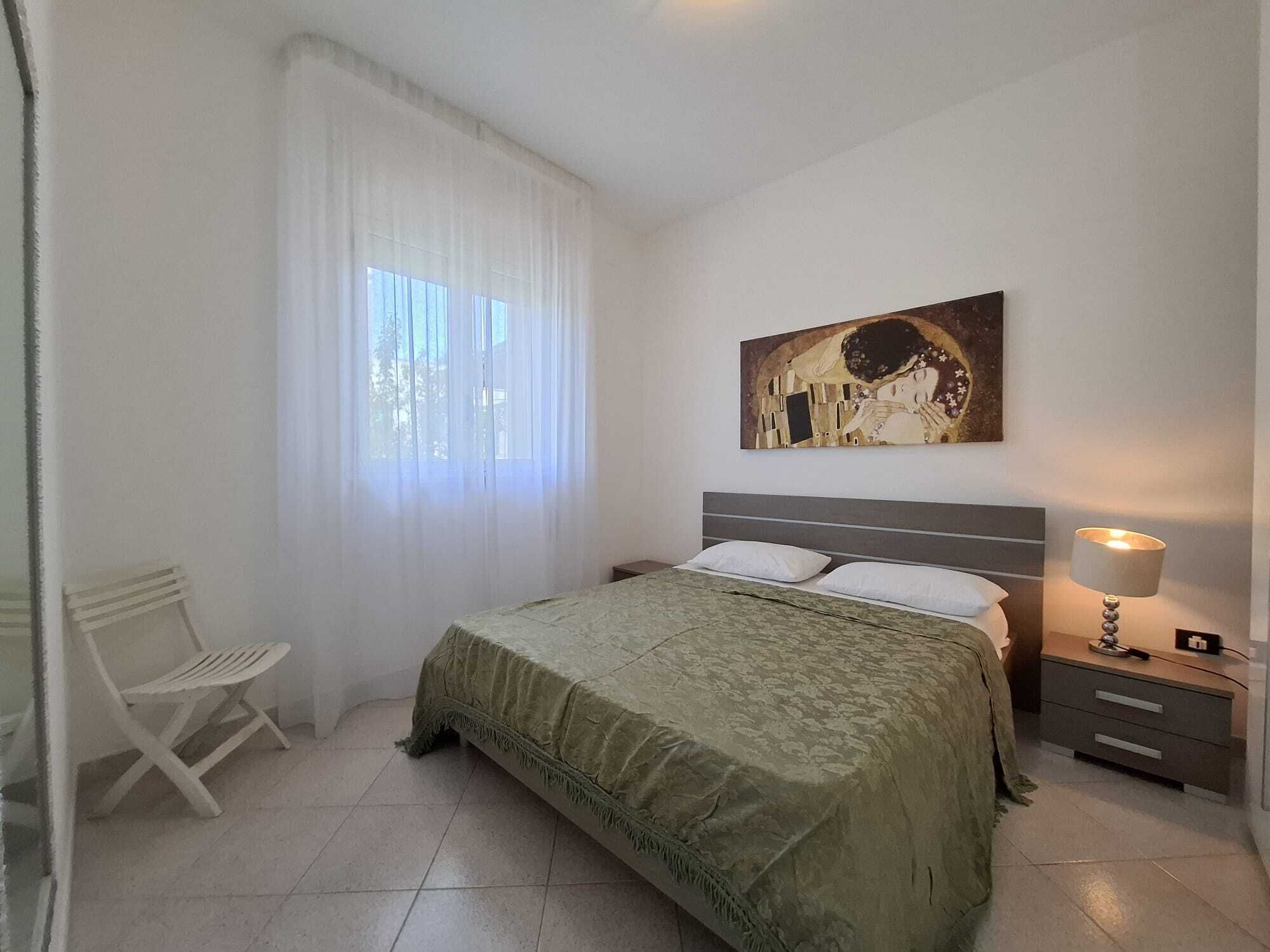 A bedroom with a bed, a chair, a bedside table with a lamp, and a painting on the wall.