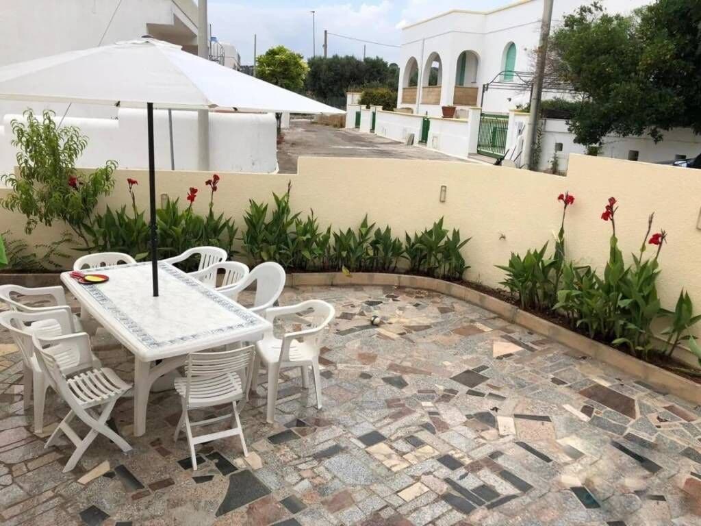 Une terrasse extérieure avec un sol carrelé, des chaises en plastique blanches et une table surmontée d’un parasol blanc.
