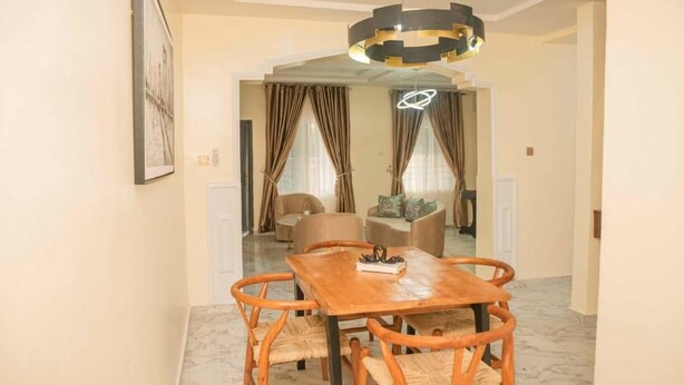 A dining area with a wooden table and wicker chairs, a chandelier, and a view into a living room with a sofa and curtains. - Tourmaline- Cozy 2-bedroom apartment in serene Lagos with AC (Lagos)