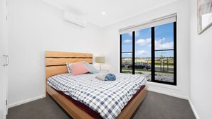 A bedroom with a wooden bed, checkered bedding, a window with a view of the street, and a ceiling air conditioner. - `Brand New`, Spacious, Breezy Comfy, Luxury Home ! (Berwick)