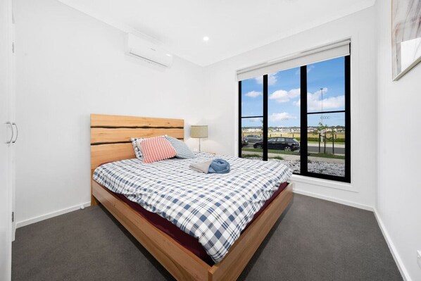 A bedroom with a wooden bed, checkered bedding, a window with a view of the street, and a ceiling air conditioner.