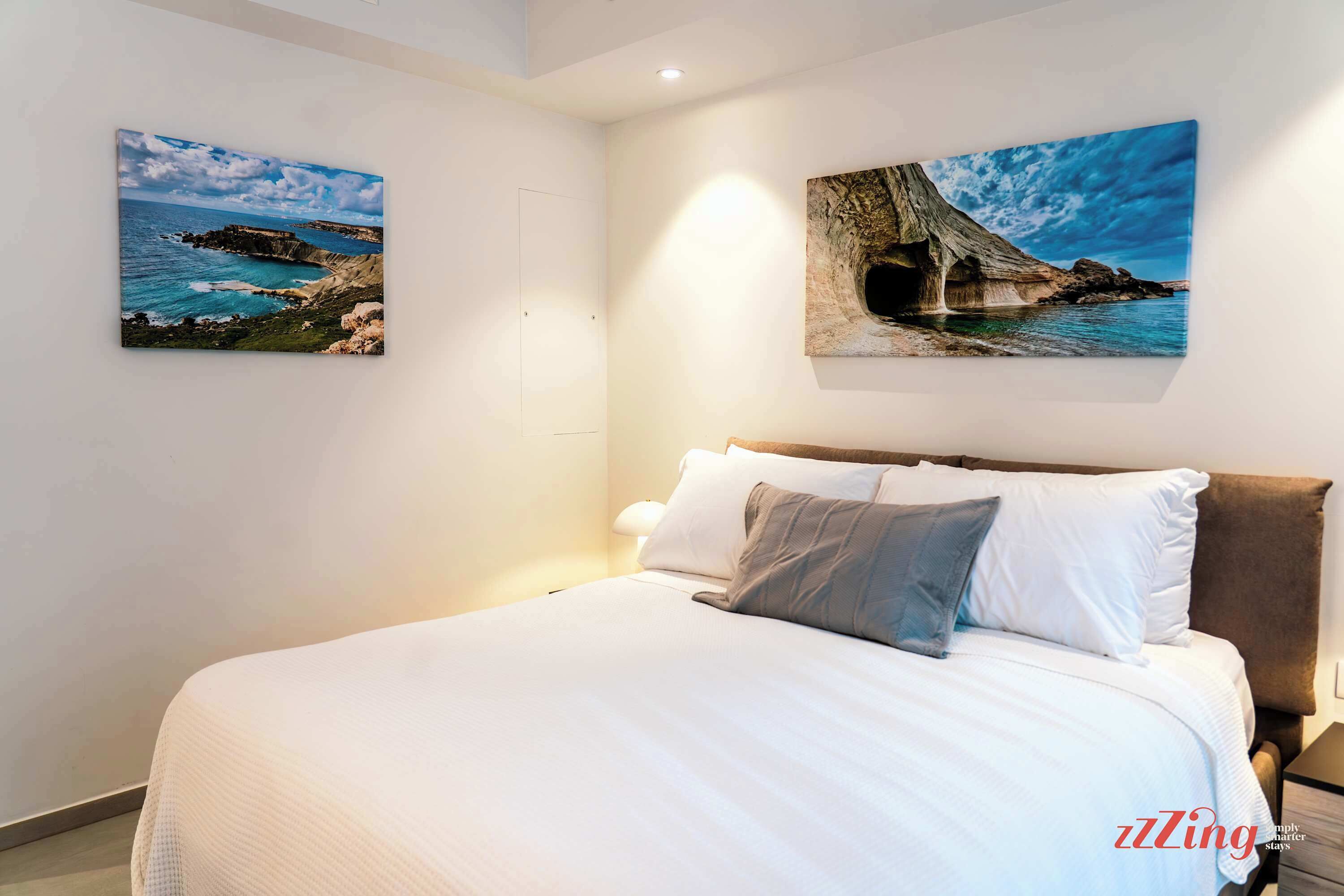 A bedroom with a large bed, two paintings of coastal scenes, and a nightstand with a lamp.